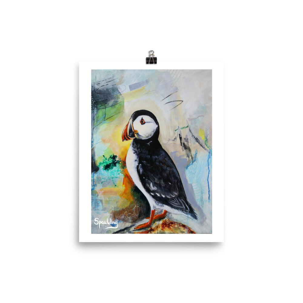 Paper art print of a black and white puffin bird with an orange beak and feet. The background colors are grey, green, yellow,blues and black.