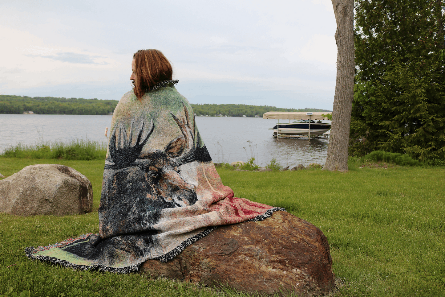 A person sits on a rock by a lake, wrapped in a blanket featuring a large moose design. The grassy area is serene, with a boat in the distance.
