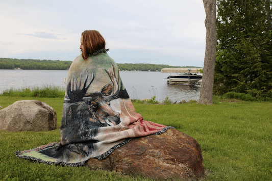 A person sits on a rock by a lake, wrapped in a blanket featuring a large moose design. The grassy area is serene, with a boat in the distance.