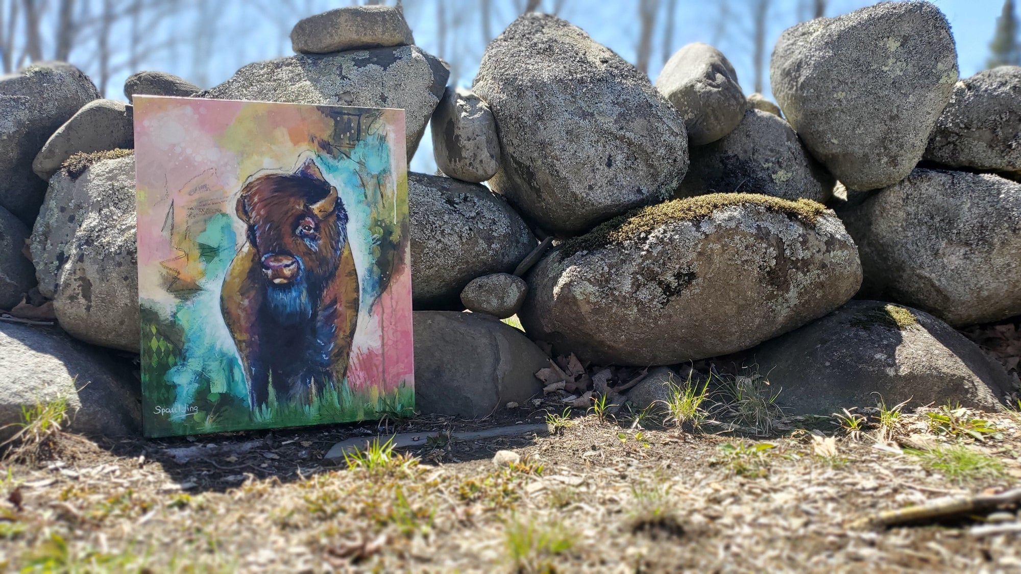 A colorful painting of a bison leans against a mossy stone wall. The bright, abstract background contrasts with the natural, rugged setting.