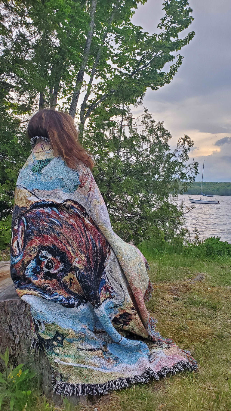 A person sits on a lakeside log, wrapped in a colorful, artistic blanket. Green trees surround the scene; a calm, overcast sky sets a peaceful ambiance.