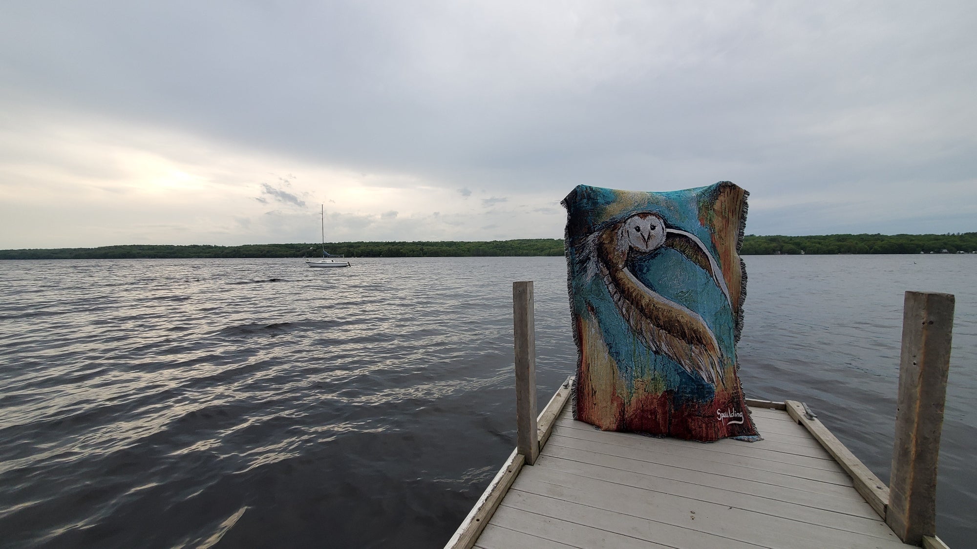 A colorful blanket with an owl design is draped over the end of a dock overlooking a calm lake. The sky is overcast, adding a serene mood to the scene.