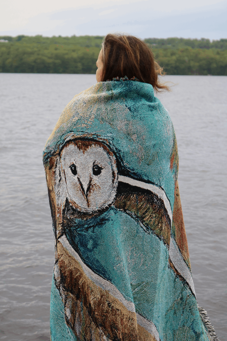 A person with long hair stands by a lake, wrapped in a blanket featuring a large owl design. The scene conveys a sense of calm and nature.