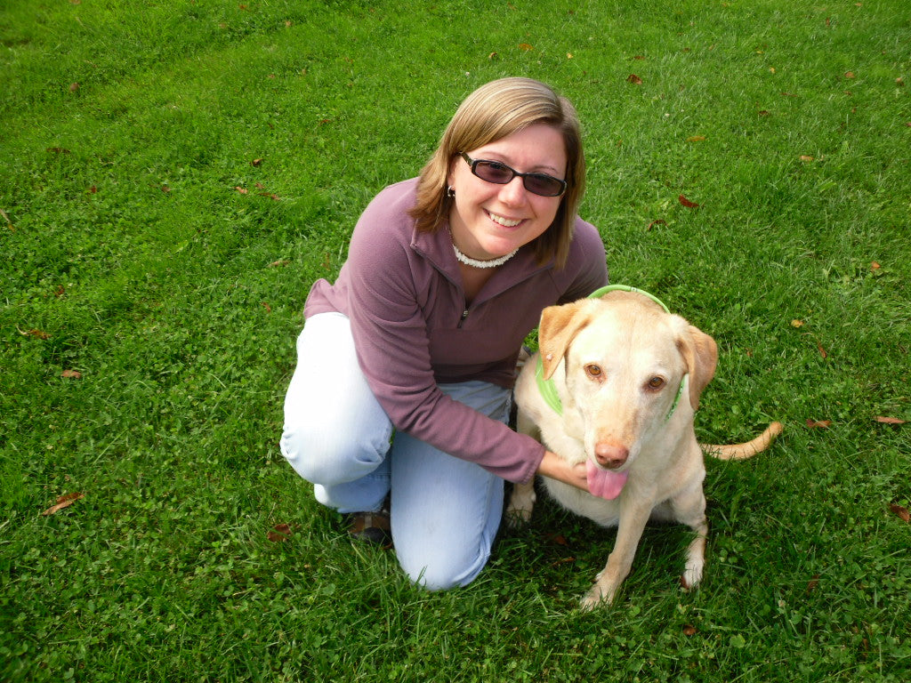A smiling woman kneels on green grass beside a tan Labrador with a bandana. The scene is cheerful and outdoors, showcasing a joyful companionship.