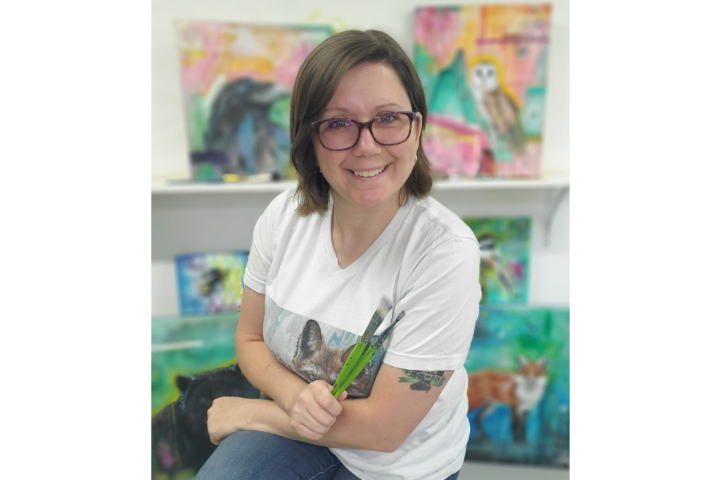 A smiling woman with glasses holds paintbrushes in a colorful art studio. She wears a white T-shirt and sits in front of vibrant animal paintings.