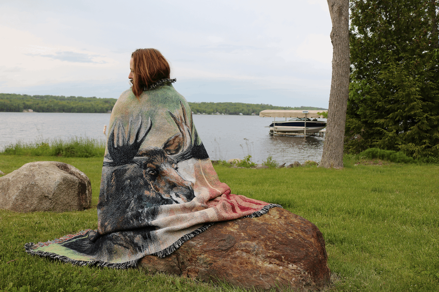 A person sits on a rock by a lake, wrapped in a blanket featuring a large moose design. The grassy area is serene, with a boat in the distance.