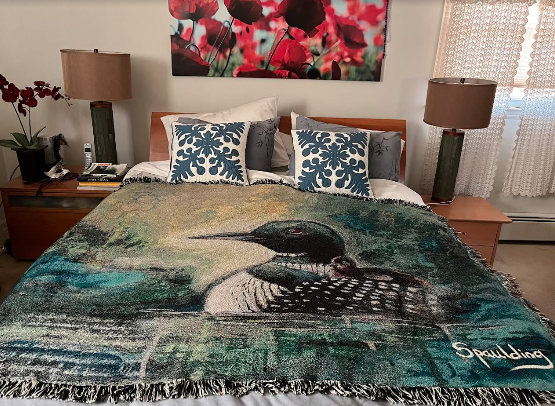 A cozy bedroom features a bed with a throw depicting a loon in water, against cushions with blue floral patterns. A vibrant poppy artwork hangs above.